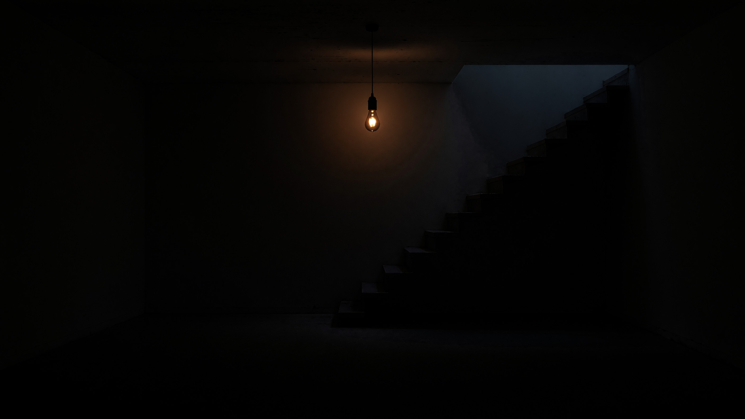 A dark empty basement: one weak bulb under the ceiling and a staircase along the back wall